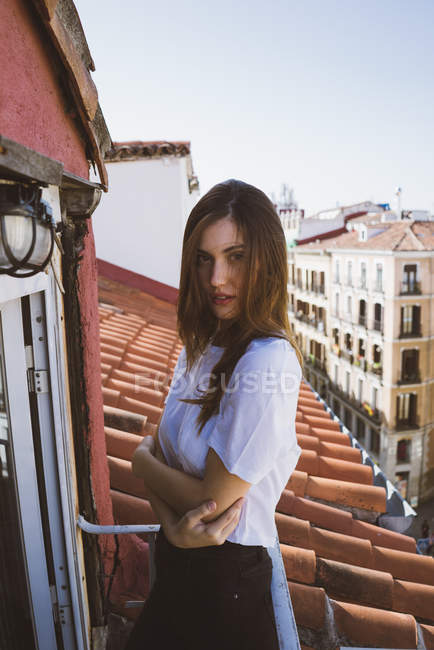 Bruna ragazza sul balcone abbracciando se stessa e guardando la fotocamera sullo sfondo di tetti della città — Foto stock