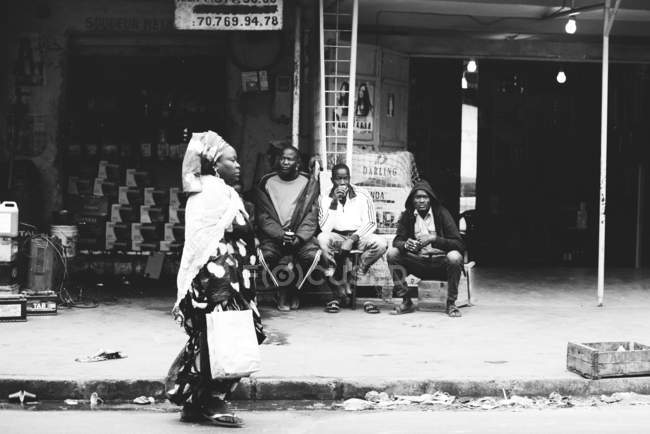 Yoff, Senegal- 6 de diciembre de 2017: Vista lateral de la mujer con bolsa de papel caminando cerca de tres hombres sentados en el banco en la escena de la calle - foto de stock