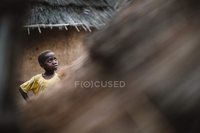 Kedougou, Senegal- 7 dicembre 2017: Ritratto oscurato di ragazzo in piedi sullo sfondo di un bungalow di paglia nel villaggio . — Foto stock