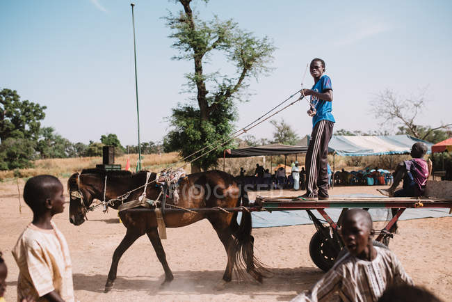 Goree, Senegal- 6 de diciembre de 2017: Vista lateral de niños africanos montando carro con caballo en el distrito rural . - foto de stock