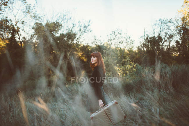 Вид сбоку рыжеволосой девушки, позирующей с чемоданом в сельской местности и романтически отводящей взгляд . — стоковое фото