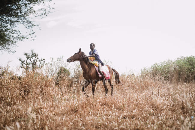 Goree, Senegal - 6 de diciembre de 2017: Niño africano cabalgando a caballo en hierba seca del campo rural . - foto de stock