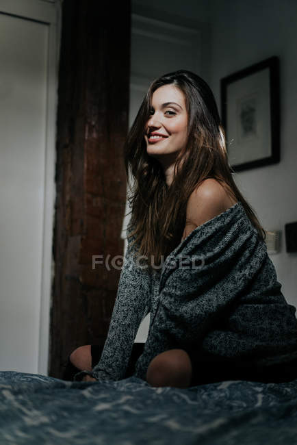 Seducente ragazza bruna seduta sul letto e sorridente alla macchina fotografica — Foto stock