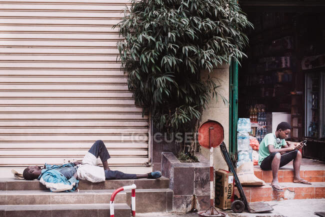 Fatigué homme africain dormir près de l'adolescent en utilisant son smartphone sur les marches de la boutique. — Photo de stock