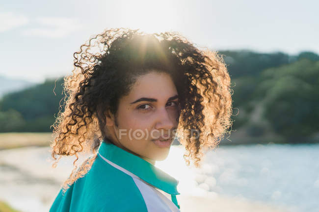 Portrait de jeune femme aux cheveux bouclés regardant au-dessus de l'épaule à la caméra sur fond de nature et de soleil éclatant . — Photo de stock
