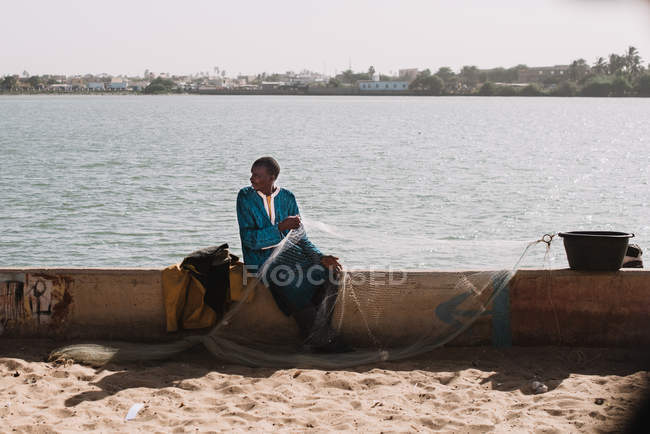 Goree, Sénégal- 6 décembre 2017 : Un homme assis avec un filet de pêche sur une clôture en béton à la lumière du soleil
. — Photo de stock