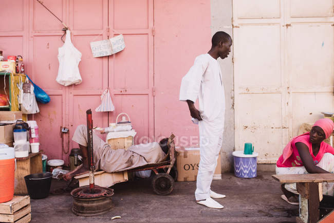 Goree, Senegal- December 6, 2017:African man in white clothes speaking to woman while standing near sleeping man. — Stock Photo