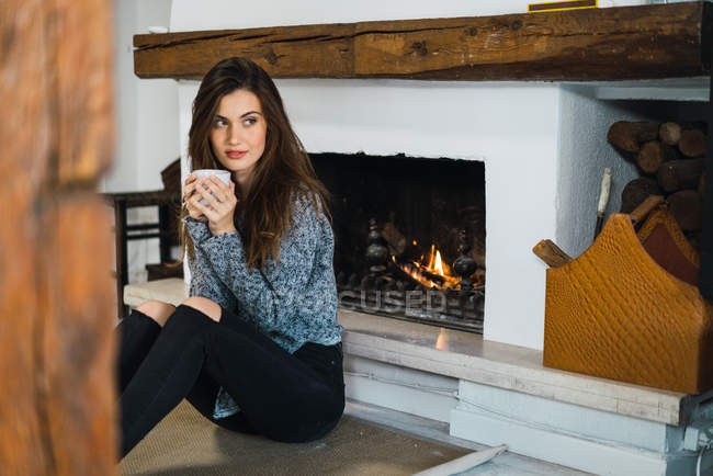 Portrait de fille brune assise avec une tasse de café par la cheminée et regardant ailleurs — Photo de stock