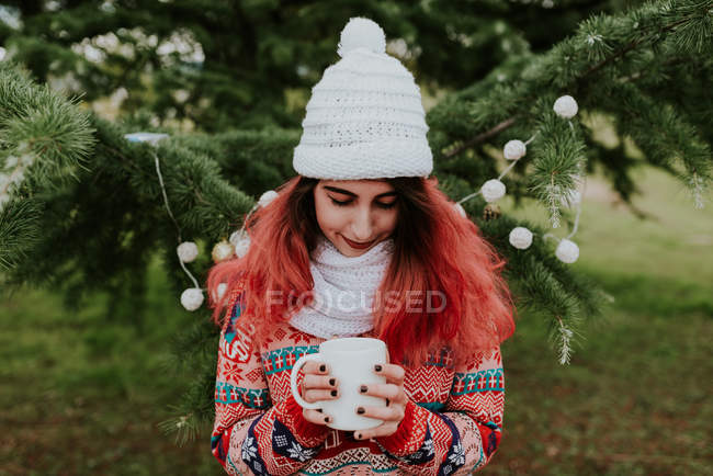 Portrait de fille en bonnet tricoté debout près du sapin festif et regardant vers le bas à la tasse avec boisson chaude dans les mains — Photo de stock