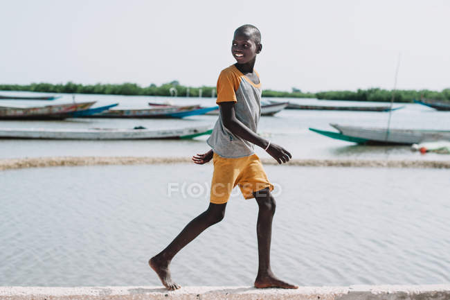 Goree, Senegal - 6 de diciembre de 2017: Sonriente niño africano mirando hacia atrás y caminando a la orilla del mar . - foto de stock