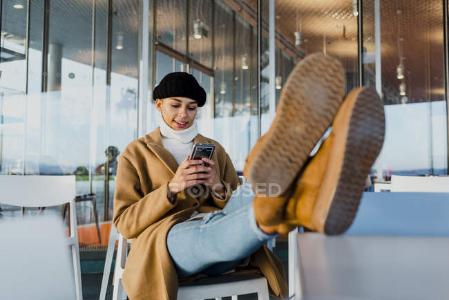 Jovem mulher em roupas quentes com os pés na mesa e usando smartphone enquanto está sentado no café . — Fotografia de Stock
