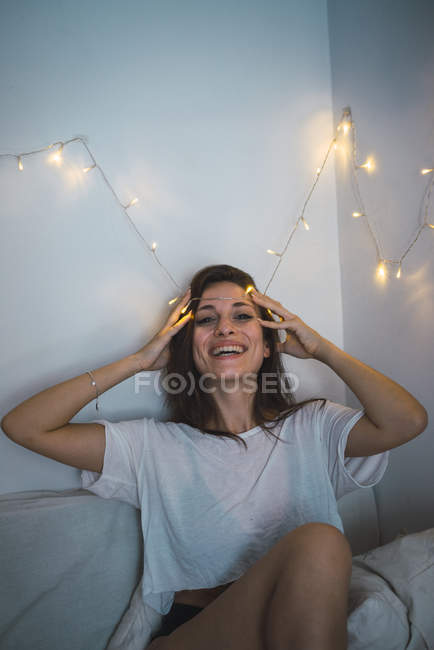 Pretty brunette girl sitting on bed and putting fairy lights on forehead. — Stock Photo