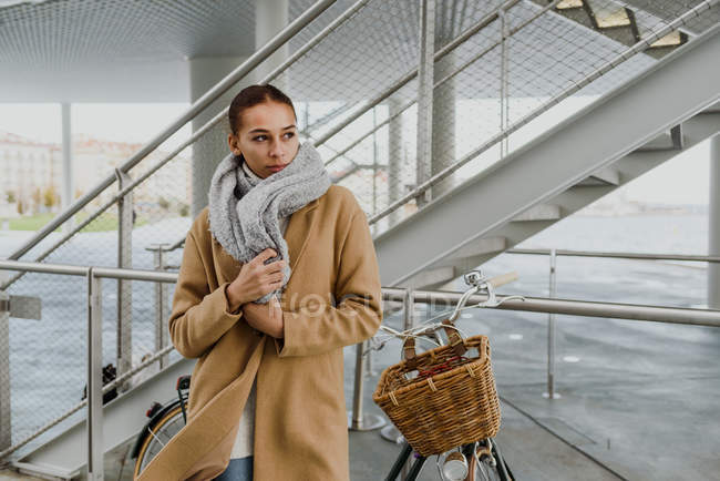 Portrait of young woman wrapping in coat and leaning on bicycle in front of staircase. — Stock Photo