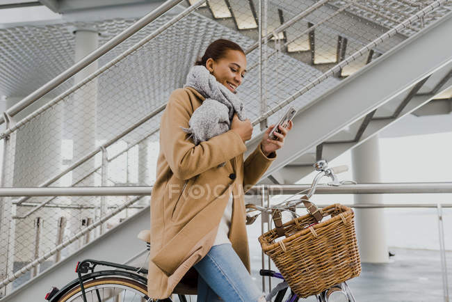 Vue latérale de la femme sur le vélo enveloppant dans un manteau et utilisant un smartphone — Photo de stock
