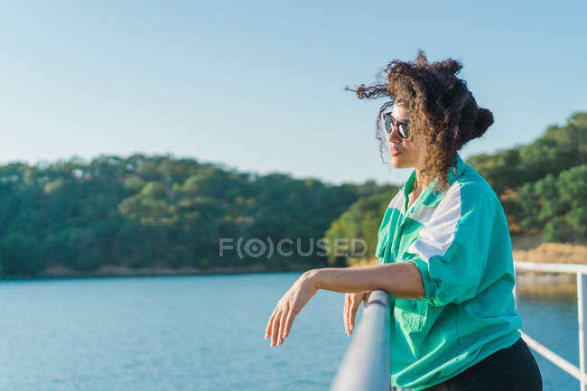 Vista lateral da mulher em óculos de sol apoiados no píer corrimão sobre lago iluminado pelo sol — Fotografia de Stock