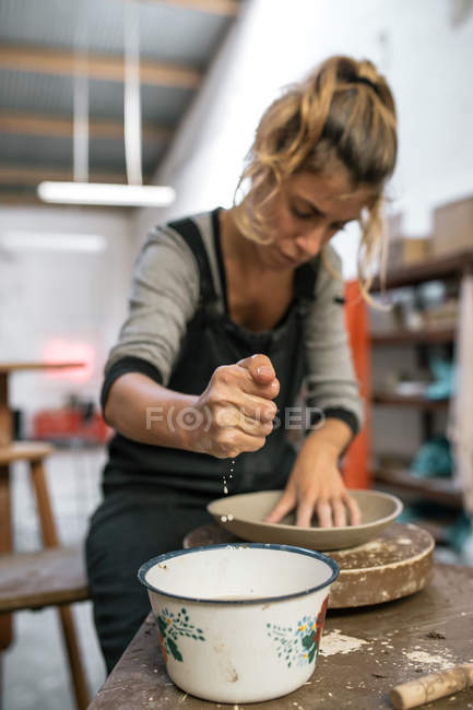 Retrato de alfarero hembra humedeciendo pieza de arcilla mientras modelaba la placa en el taller - foto de stock