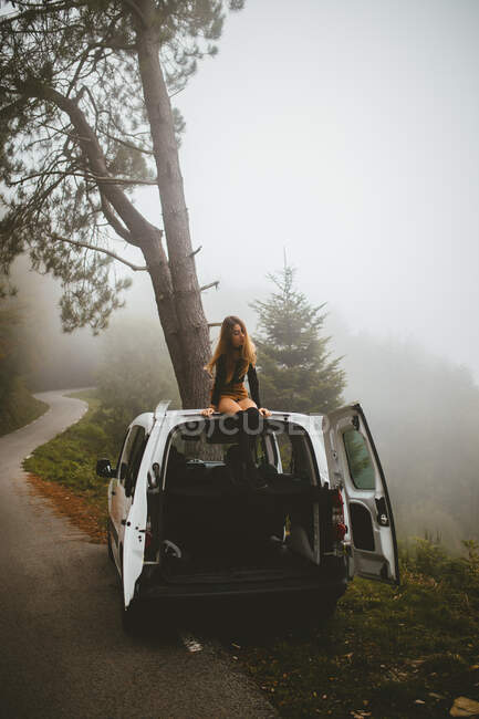 Гарненька молода жінка сидить на вершині автомобіля під час паркування на узбіччі сільської дороги . — стокове фото
