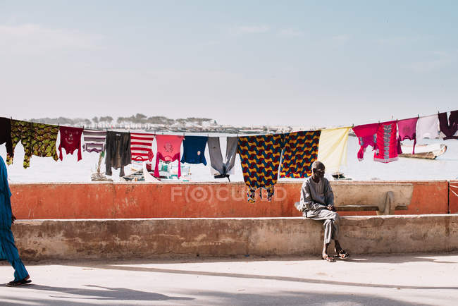 Goree, Senegal - 6 de diciembre de 2017: Vista del hombre sentado en la calle de detrás de la ropa de cama en la cuerda a la luz del sol . - foto de stock