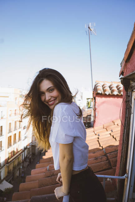 Side view of smiling brunette girl posing on balcony over rooftops cityscape and looking at camera — Stock Photo
