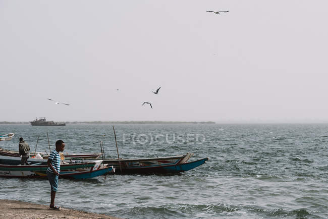 Goree, Sénégal- 6 décembre 2017 : Vue des gens sur la côte avec de vieux bateaux flottant sur l'eau et des oiseaux volant . — Photo de stock