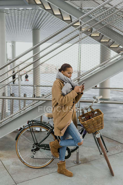 Pretty woman with city bicycle wrapping in coat and browsing smartphone — Stock Photo