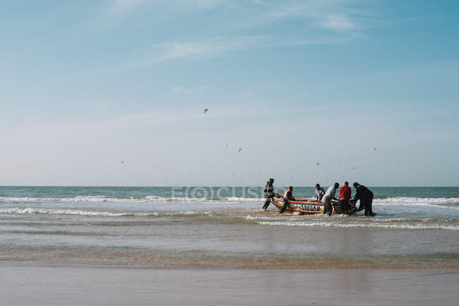 Yoff, Senegal - December 6, 2017: Distant view to group of people at beach pushing boat to ocean — стоковое фото