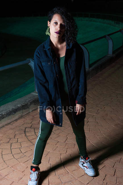 Портрет девушки в городской одежде, позирующей на ночной улице и смотрящей в камеру — стоковое фото