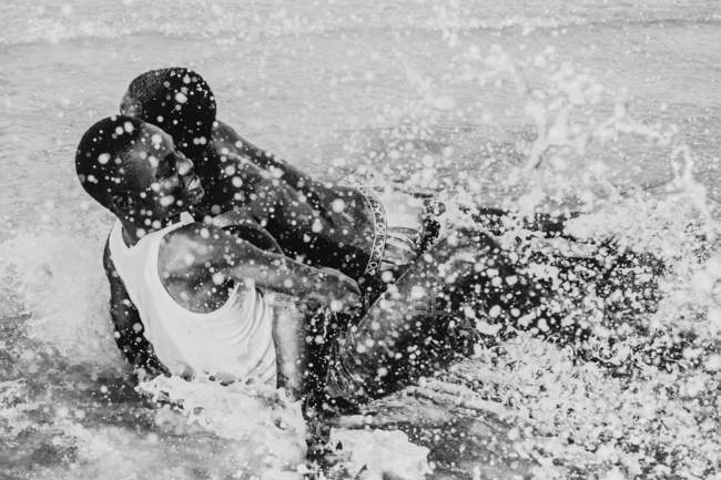 Yoff, Senegal- December 6, 2017: Young men wrestling cheerfully in shallow water of ocean shoreline. — Stock Photo