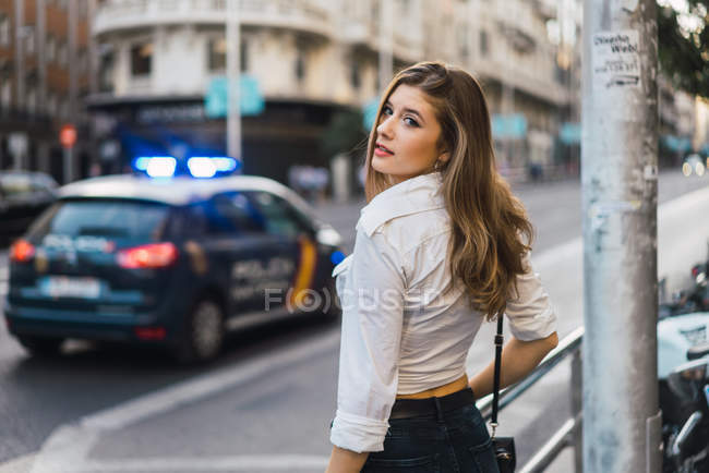 Menina morena posando na cena da rua com carro de polícia borrado e olhando sobre o ombro na câmera — Fotografia de Stock