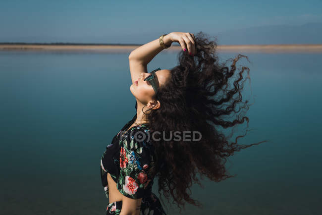 Vista lateral da menina morena em óculos de sol acenando com o cabelo sobre a paisagem do lago — Fotografia de Stock