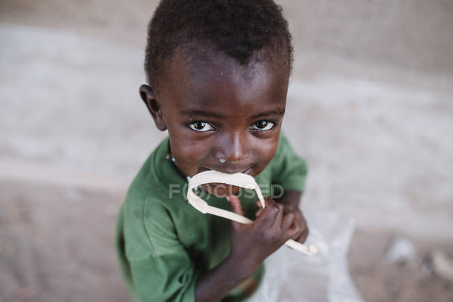 Goree, Sénégal- 6 décembre 2017 : Portrait d'un petit garçon noir mordant un ruban en plastique sur la scène de rue et regardant une caméra — Photo de stock