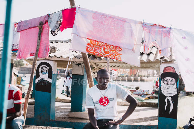 Goree, Senegal- Dezembro 6, 2017: Retrato de homem sentado sob roupa de secagem e olhando para a câmera . — Fotografia de Stock