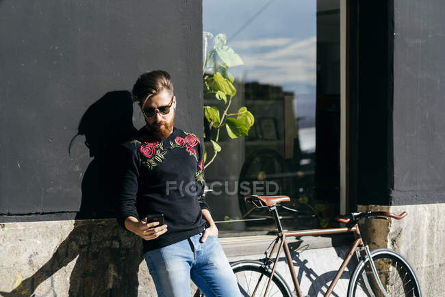 Uomo alla moda appoggiato al muro in bicicletta e smartphone di navigazione — Foto stock