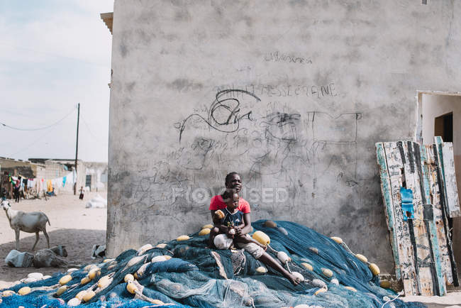Goree, Senegal- December 6, 2017: Woman with child sitting on huge fishing net near concrete wall at street scene. — Stock Photo