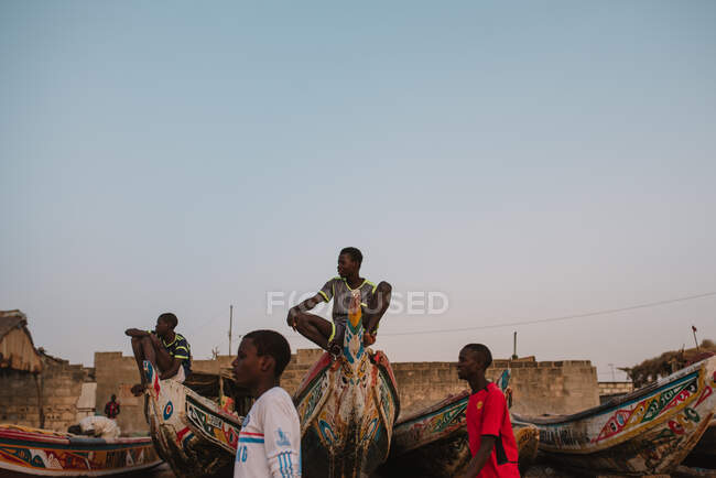 African teenagers walking on beach near boats and sitting on them. — Stock Photo