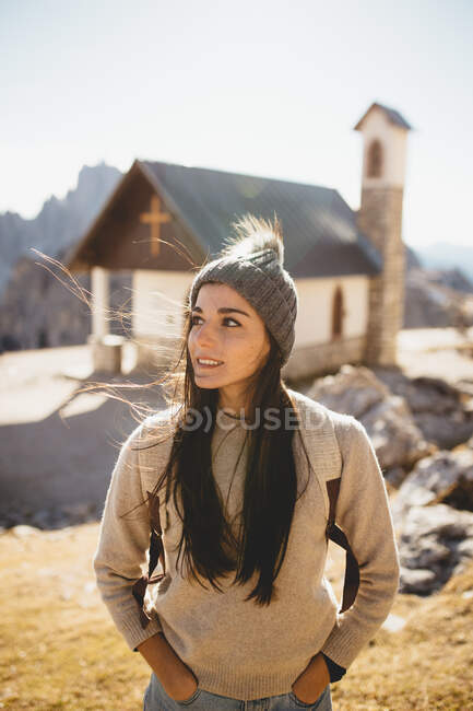 Pretty young woman in hat standing near small church and enjoying weather on sunny day. — Stock Photo