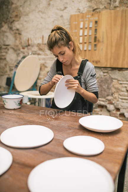 Mulher concentrada em avental sentado à mesa e formando placas de barro branco . — Fotografia de Stock