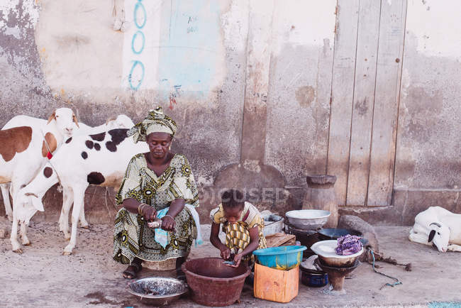 Goree, Sénégal- 6 décembre 2017 : Femme africaine et sa fille assise dans la rue et coupant des légumes sur fond de chèvres par mur de béton . — Photo de stock