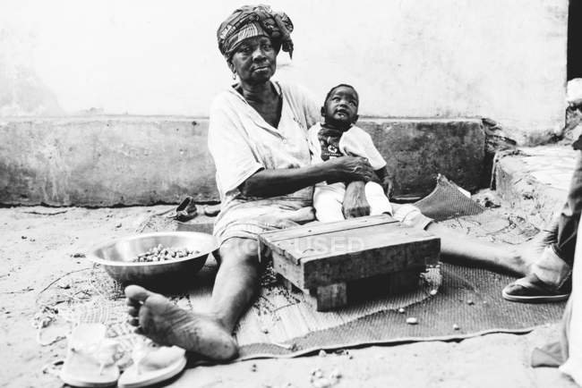 Yoff, senegal- 6. Dezember 2017: Porträt einer barfüßigen Frau mit kleinem Jungen, die auf schmutzigem Straßenpflaster sitzt und in die Kamera blickt — Stockfoto
