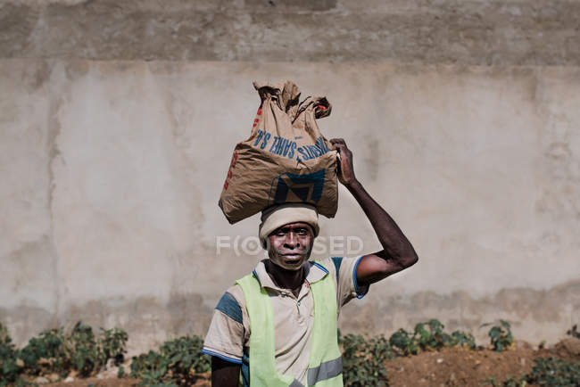 Goree, Senegal- 6 dicembre 2017: Ritratto di uomo che porta borsa di sostanza secca sulla testa guardando la macchina fotografica . — Foto stock