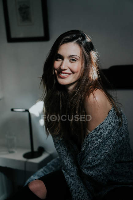 Portrait of young brunette girl sitting on bed and smiling at camera — Stock Photo