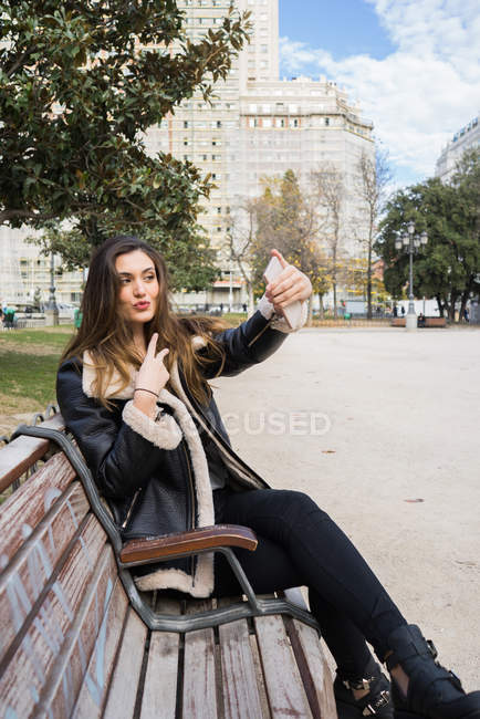 Portrait de femme brune prenant selfie avec smartphone au banc du parc — Photo de stock