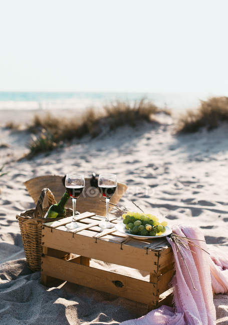Натюрморт из бокалов с вином и тарелки с белым виноградом, стоящим на деревянной коробке на пляже . — стоковое фото