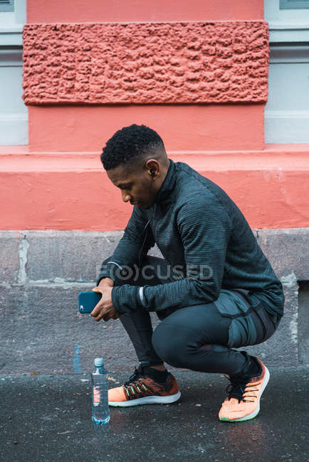 Vista lateral del atleta en cuclillas cerca de la botella de agua y sosteniendo el teléfono inteligente en las manos - foto de stock