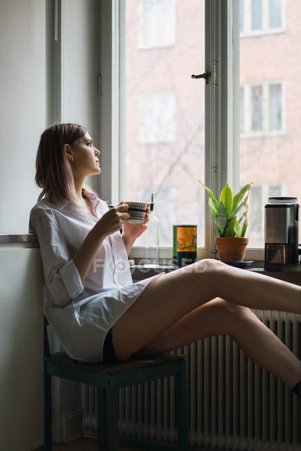 Вид сбоку женщины с чашкой, сидящей у окна и отводящей взгляд — стоковое фото