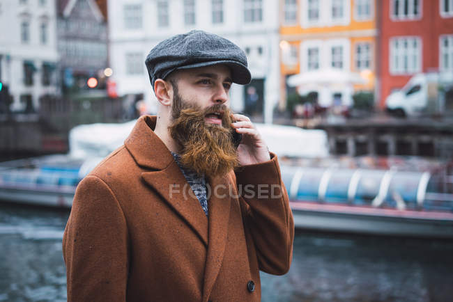 Retrato de hombre barbudo en ropa vintage hablando ensmartphone en el río en la ciudad - foto de stock
