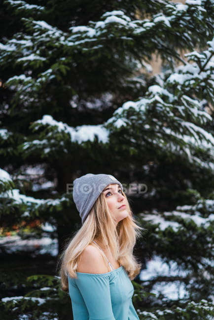 Vista lateral de la chica sensual rubia de pie en el árbol de invierno y mirando hacia arriba - foto de stock
