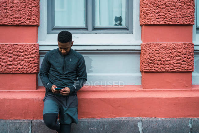 Sportive man leaning on building facade and looking down at smartphone in hands — Stock Photo