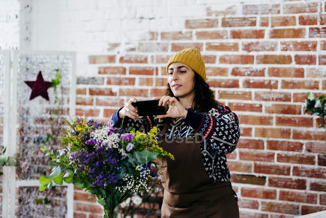 Улыбающаяся женщина в трикотажном свитере и шляпе фотографирует букет — стоковое фото