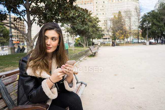 Портрет брюнетки, сидящей на скамейке в парке со смартфоном и отворачивающейся — стоковое фото